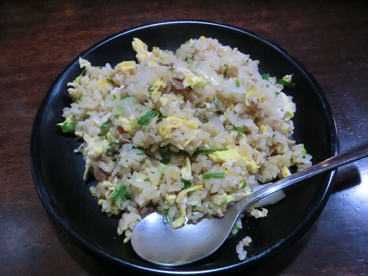 A close-up photo shows a black bowl filled with fried rice containing scrambled egg, chopped green onions, and translucent…