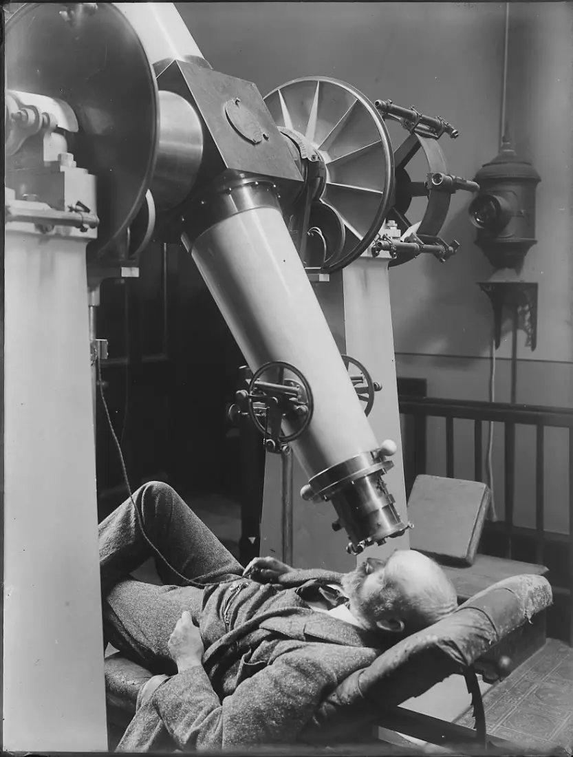 A black-and-white photograph shows an elderly man with a beard lying back on a chair, looking up through the eyepiece of a…