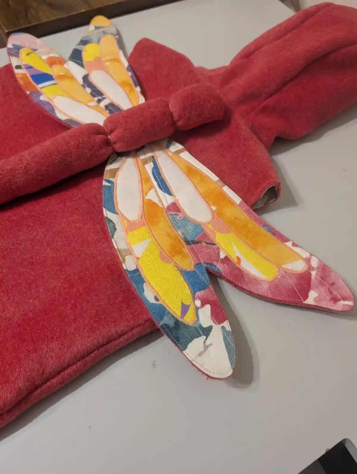 A pair of red felt oven mitts are folded and arranged to resemble a dragonfly, with colorful patterned fabric wings attached…