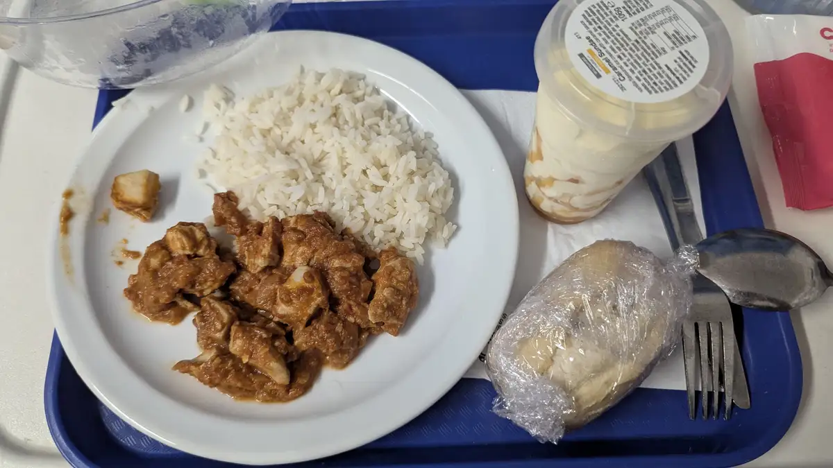 A hospital meal tray holds a plate with rice and curry, a plastic cup of caramel sundae, and a banana wrapped in plastic…