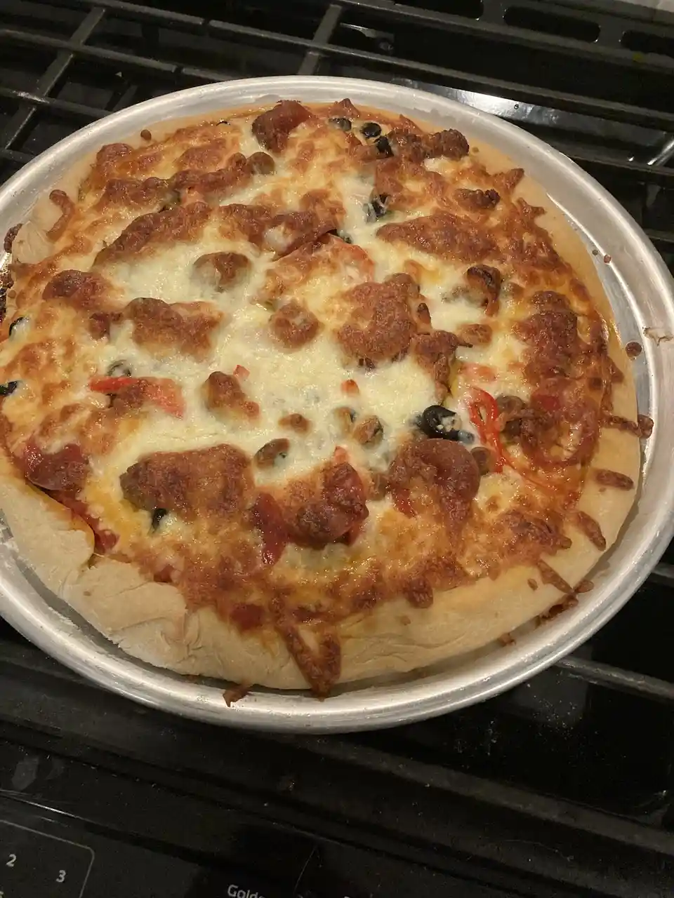 A freshly baked pizza with melted cheese, pepperoni, black olives, and red peppers sits on a metal pan atop a black…