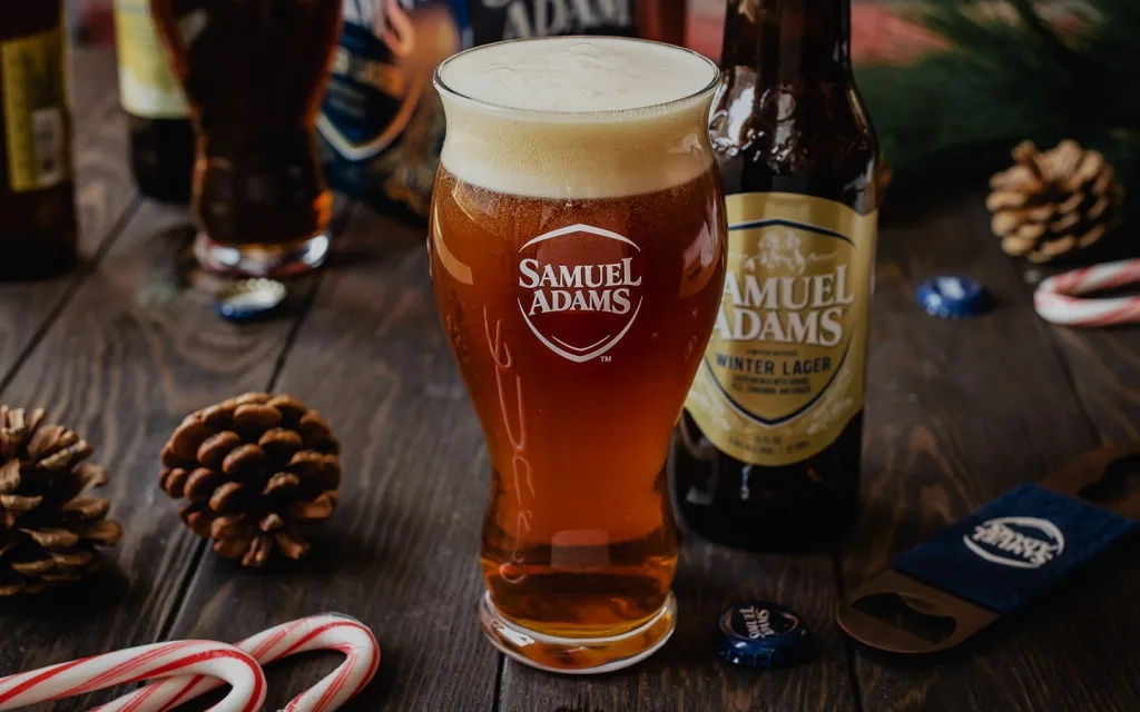 A glass of Samuel Adams Winter Lager beer with a thick white head sits on a wooden table, surrounded by pinecones, candy…