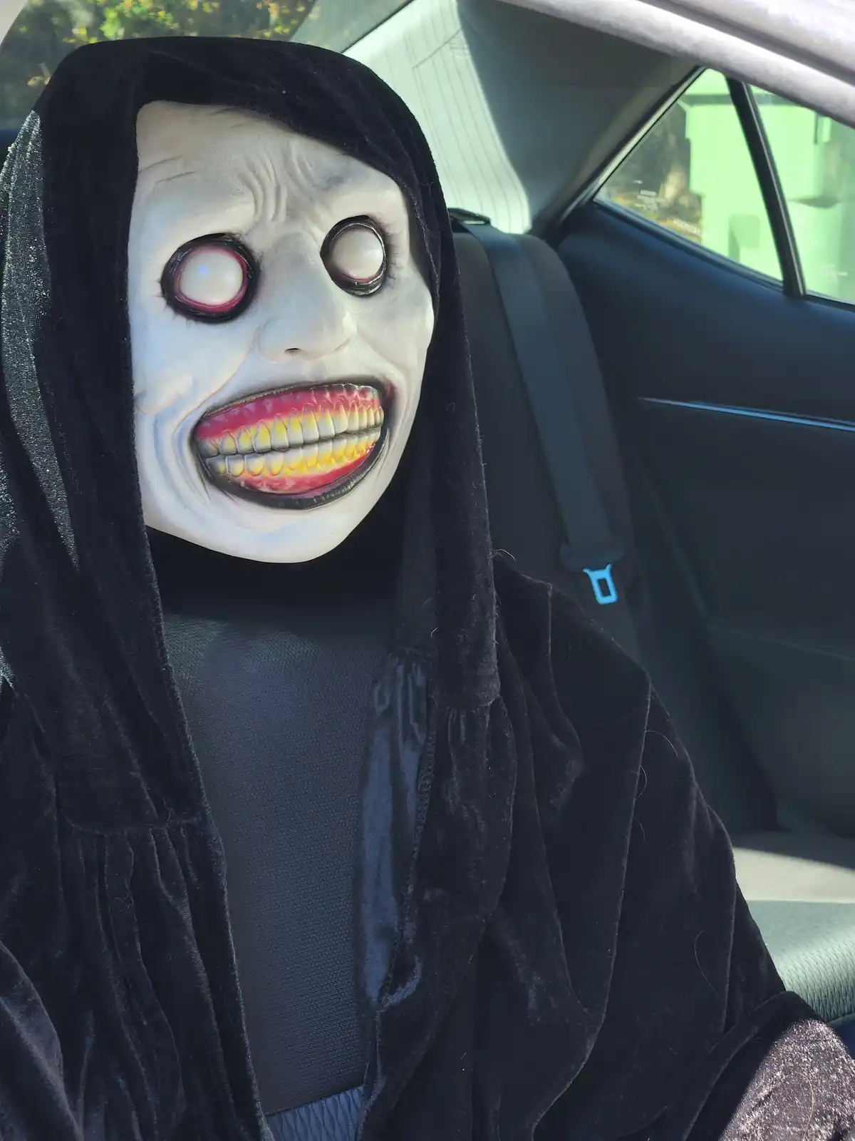 A person wearing a black hooded robe and a grotesque, wide-grinning horror mask with large white eyes sits in the passenger…