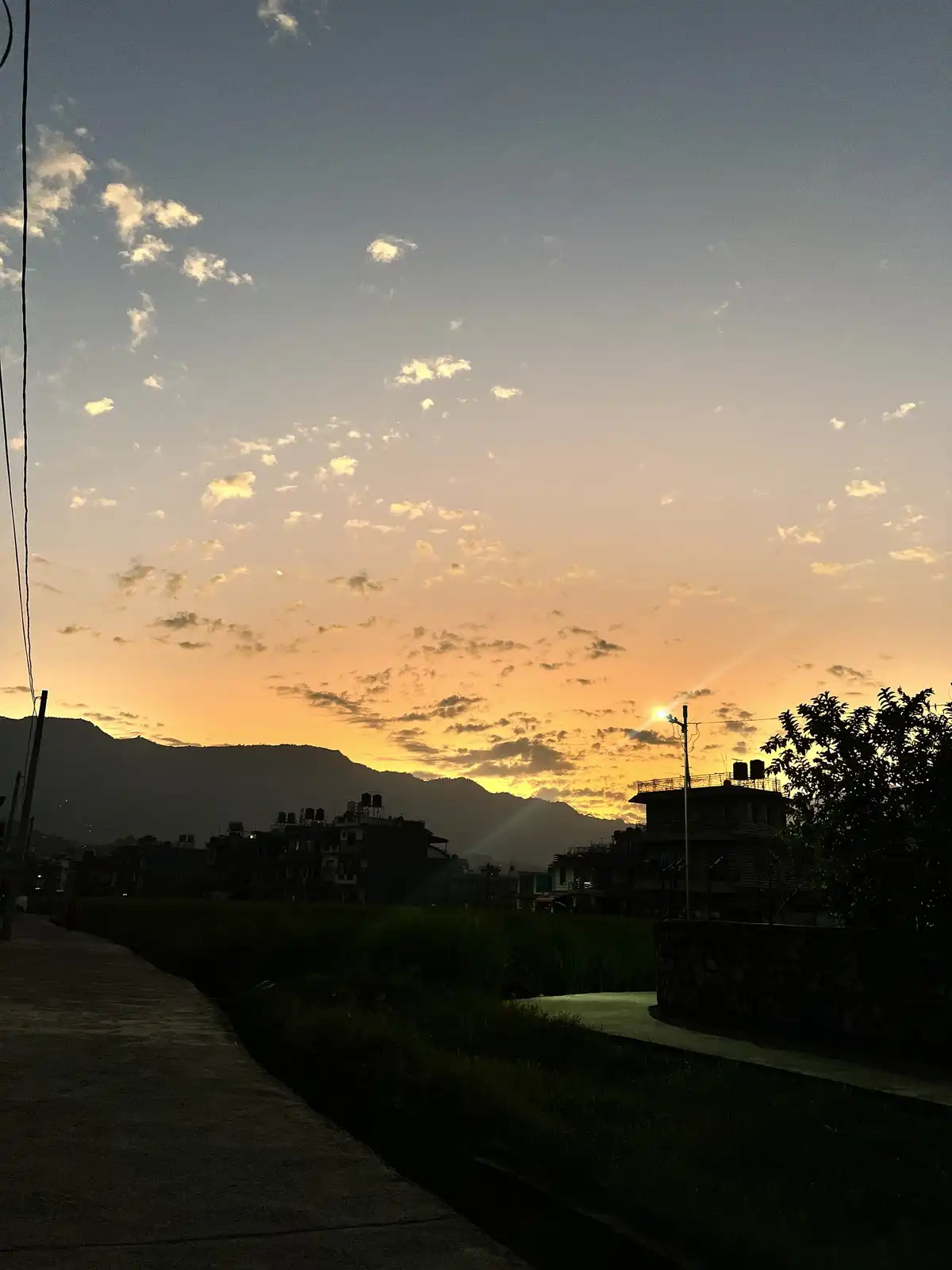 A serene sunset photo showing a rural landscape with silhouetted buildings, mountains, and a paved path in the foreground…