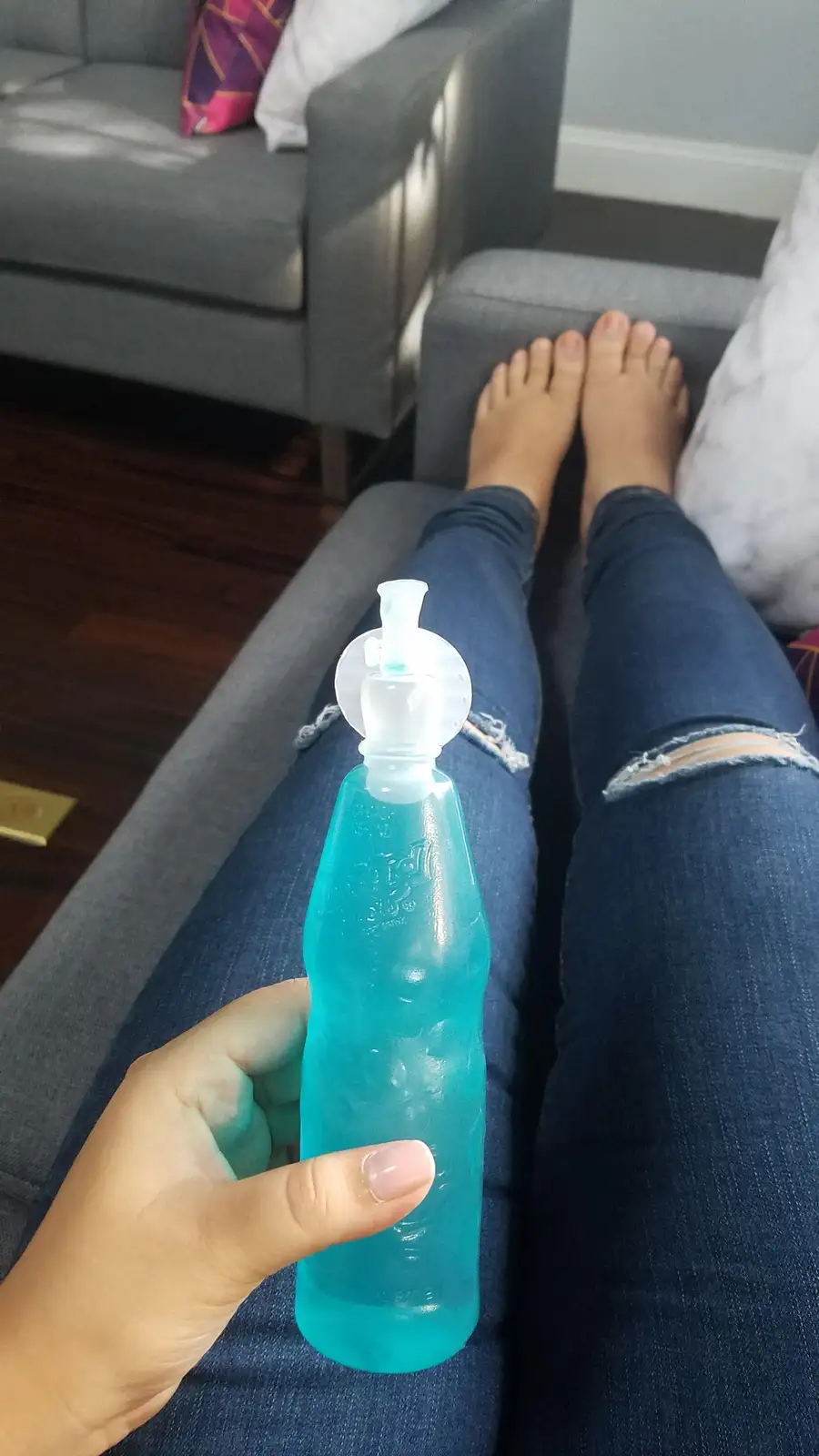 A person is sitting on a gray couch wearing ripped blue jeans, holding a bright blue bottle of dish soap with a white pump…