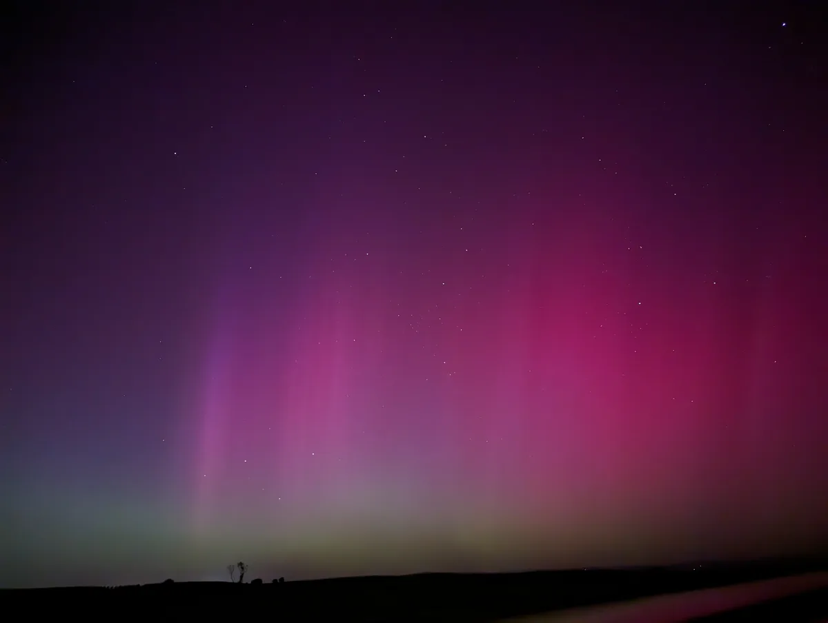 A stunning night sky photograph shows vibrant pink and purple auroras dancing above a dark, starry expanse, with a…