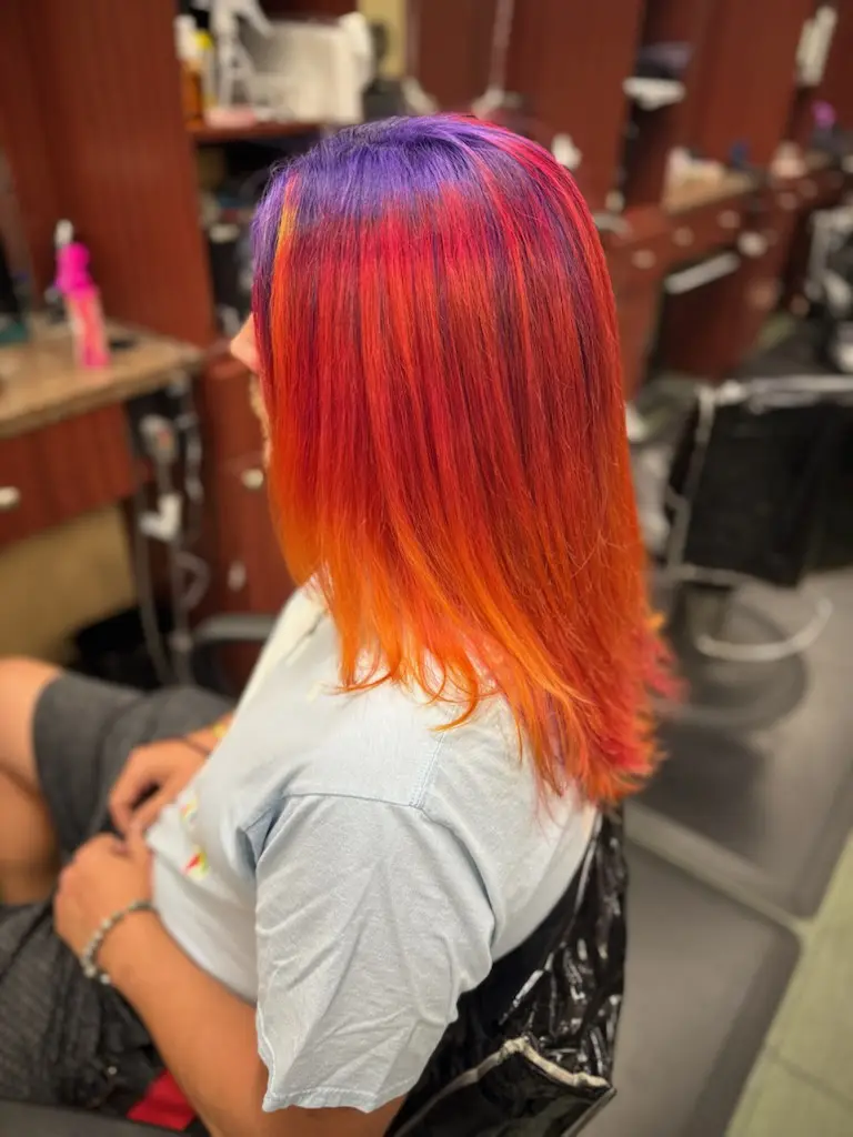 A person sits in a salon chair, viewed from behind, showcasing vibrant, multi-colored hair transitioning from purple at the…