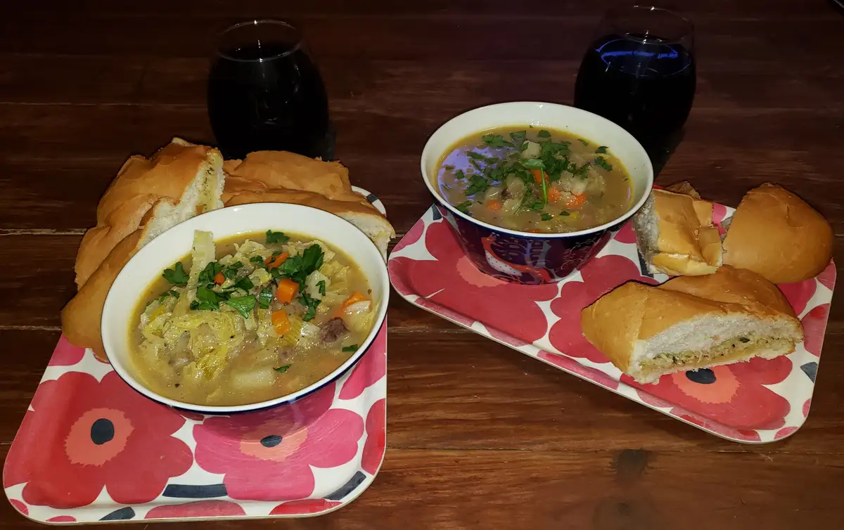 Two bowls of hearty soup, garnished with parsley and carrots, sit on floral-patterned trays with sliced bread rolls and…