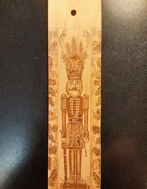 A wooden bookmark with a pyrographed nutcracker soldier wearing a crown topped with pine branches, standing between…