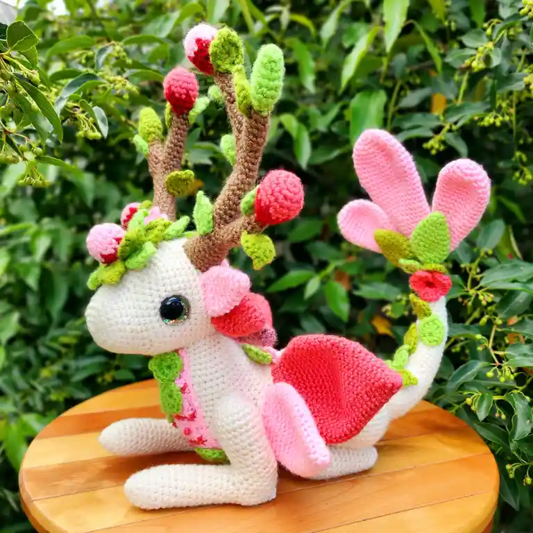 A white crocheted dragon toy sits on a wooden table outdoors, adorned with pink and green crocheted cherry blossoms and…