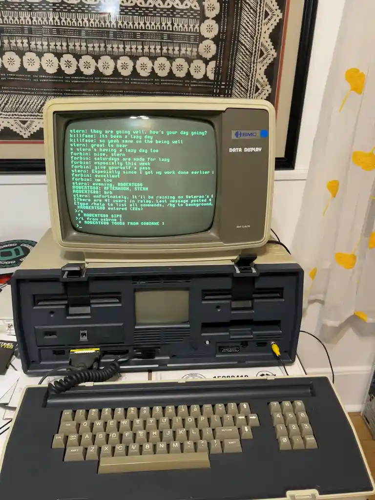 An old Osborne 1 portable computer with its screen displaying a green text-based online chat log between users like 'stern'…