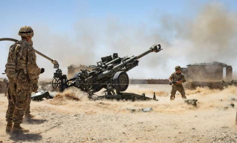 Two soldiers in desert camouflage stand beside a large, recently fired artillery piece that is kicking up dust and smoke…