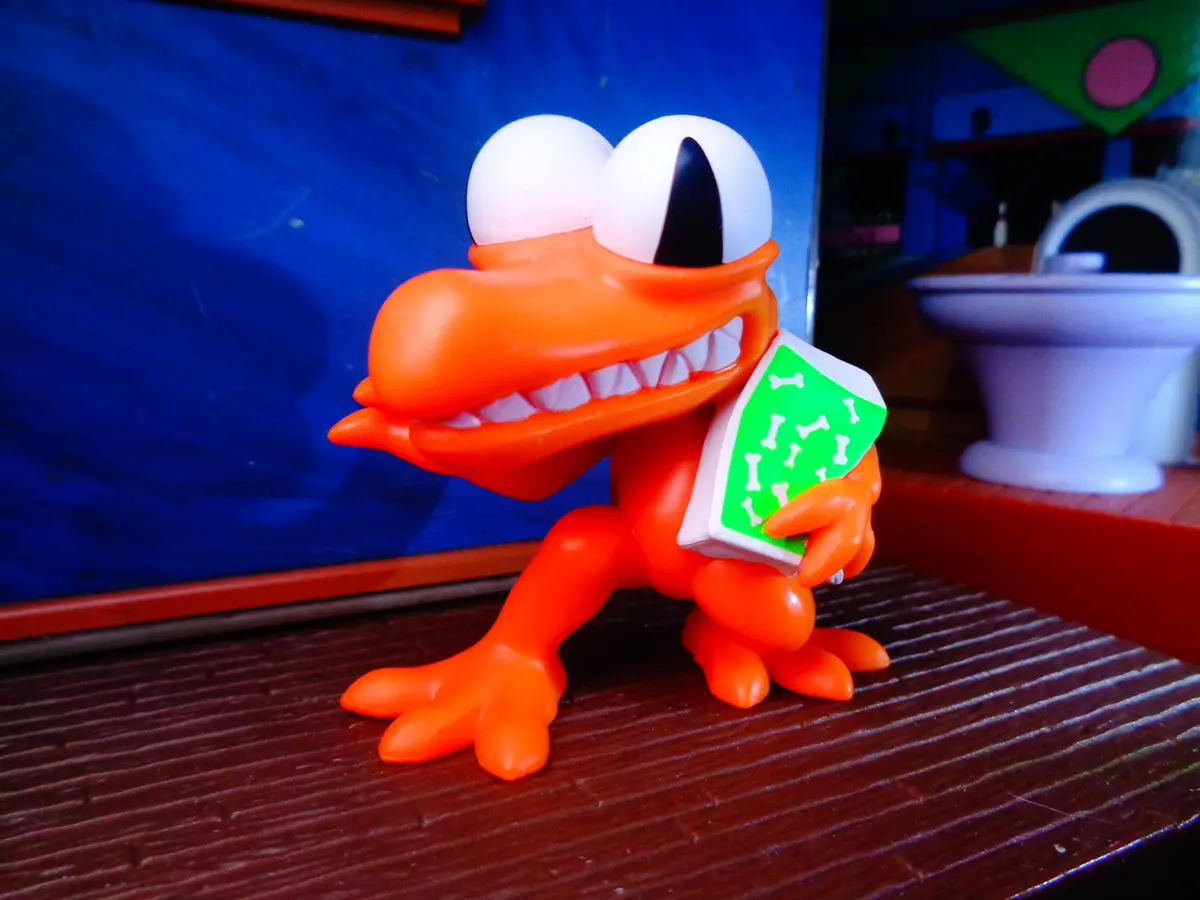 An orange plastic toy crocodile with large white eyes and a wide grin stands on a wooden surface, holding a green phone…