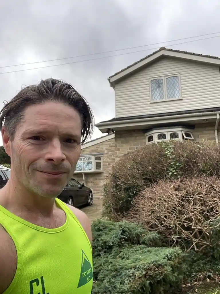 A man in a bright neon green athletic tank top stands outside a house, looking directly at the camera with a smug…