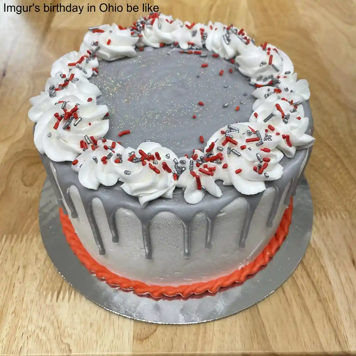 A round birthday cake with gray frosting, white piped rosettes around the top edge, and red and silver sprinkles. The cake…
