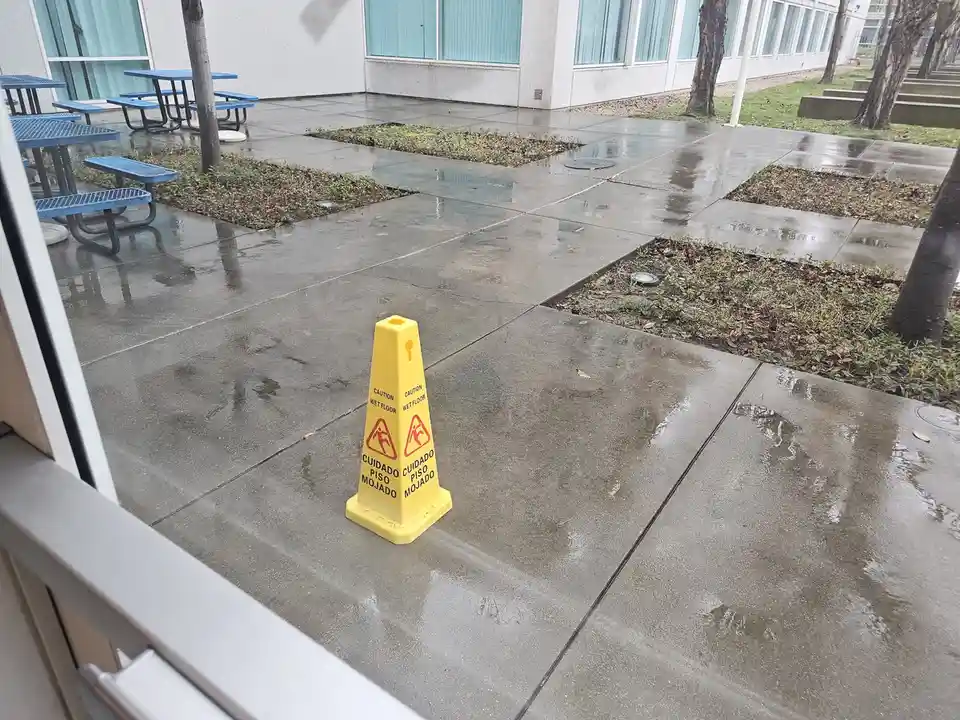 A yellow wet floor cone stands on a wet concrete patio, displaying the warning 'CAUTION WET FLOOR' in English and 'CUIDADO…