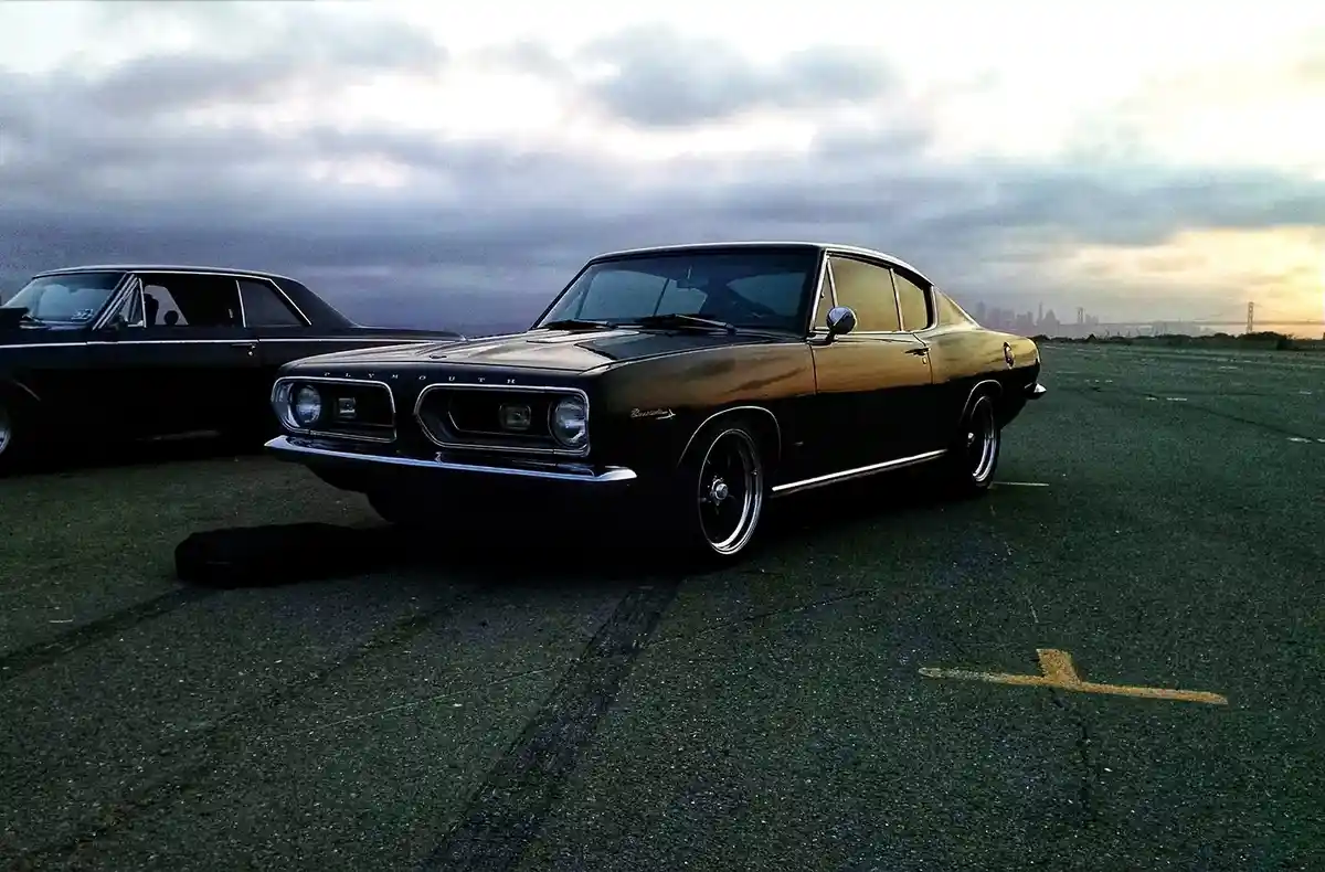 A black 1960s Plymouth Barracuda muscle car is parked on asphalt with a city skyline and bridge in the background under a…