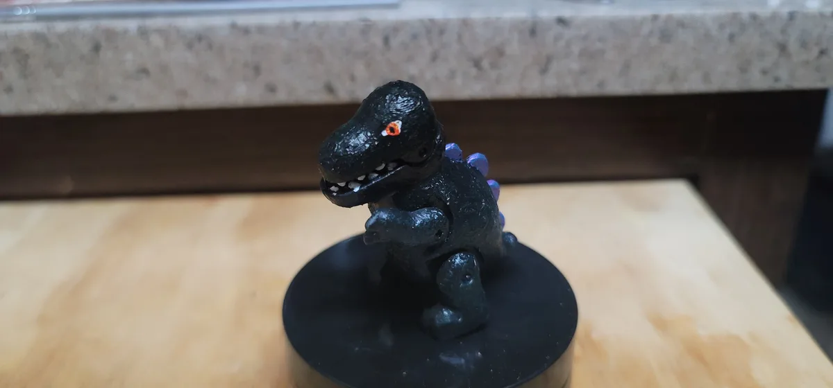 A small, black-painted toy dinosaur with glowing orange eyes and purple spikes sits on a black circular base on a wooden…