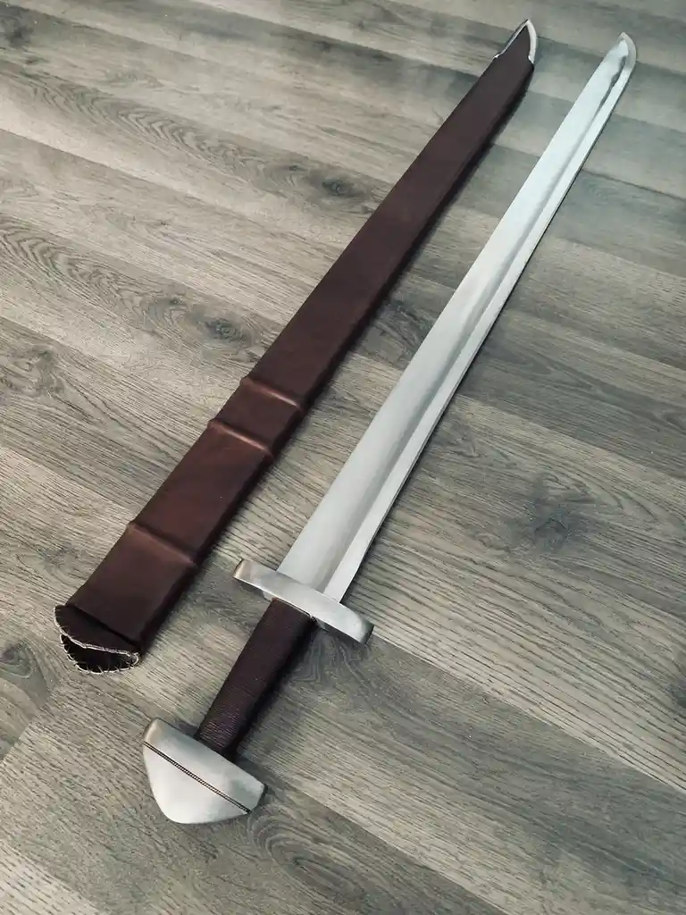A shiny, silver sword with a brown leather-wrapped hilt and pommel lies next to its matching brown leather scabbard on a…