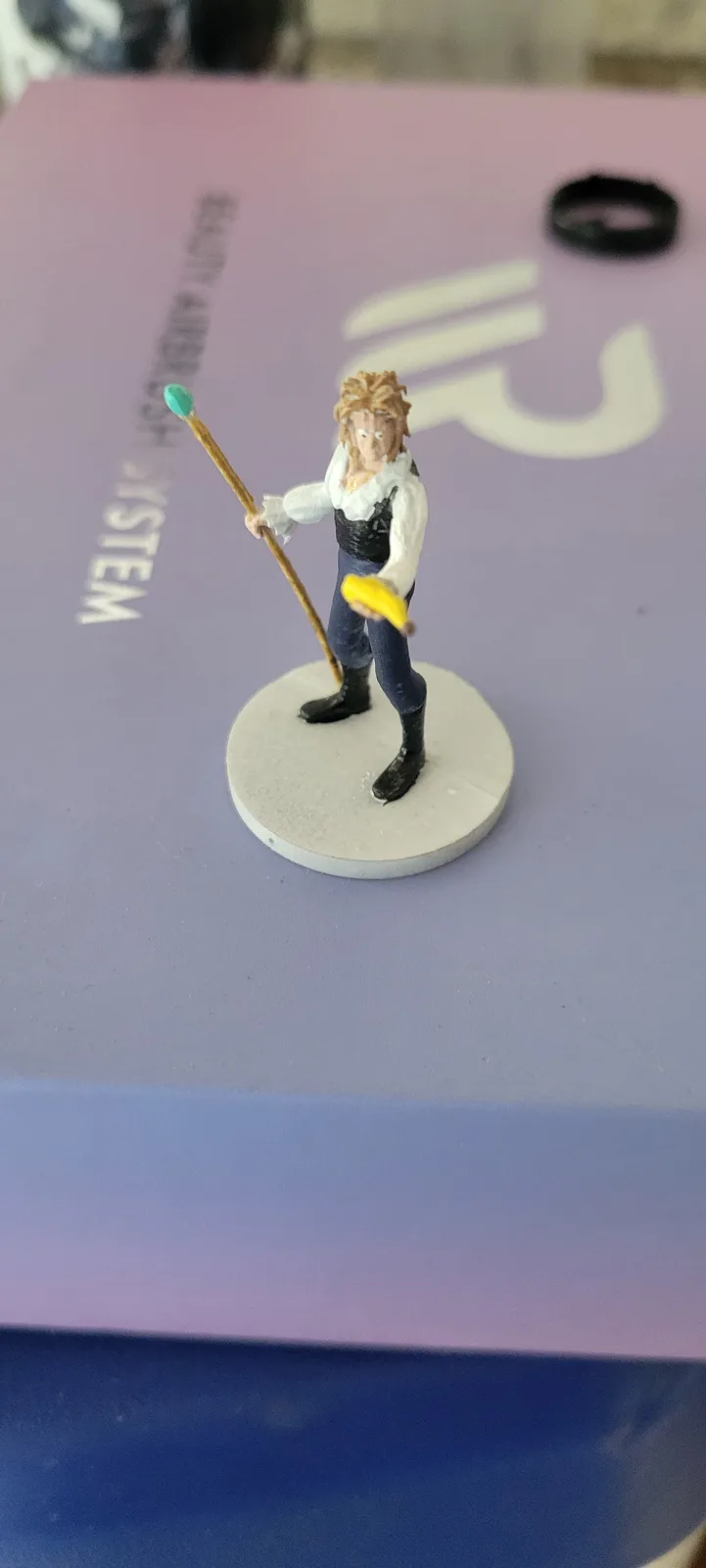 A miniature figurine of a man with spiky hair, wearing a white shirt and blue pants, stands on a circular base holding a…