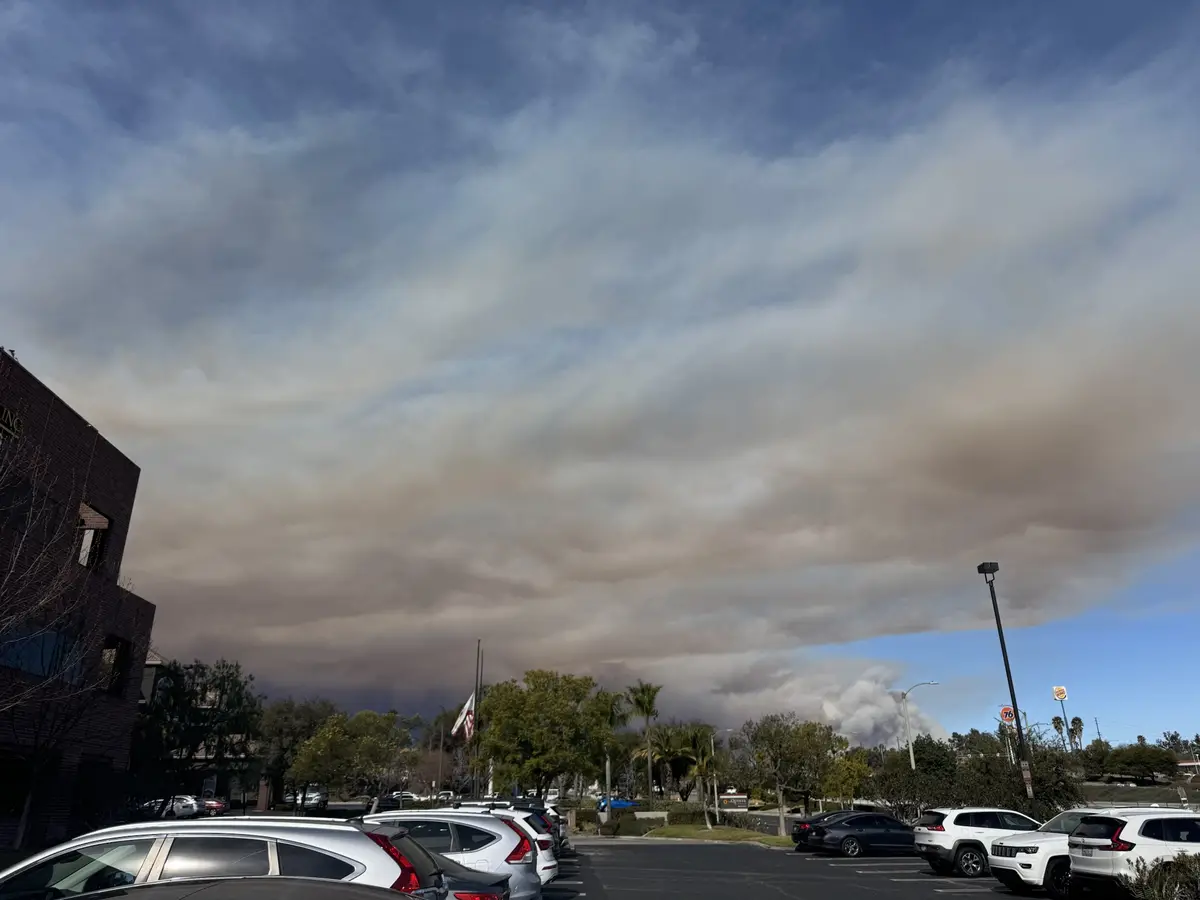 A parking lot with cars and buildings under a sky dominated by thick, brownish-gray smoke that looks like a cloud, with a…