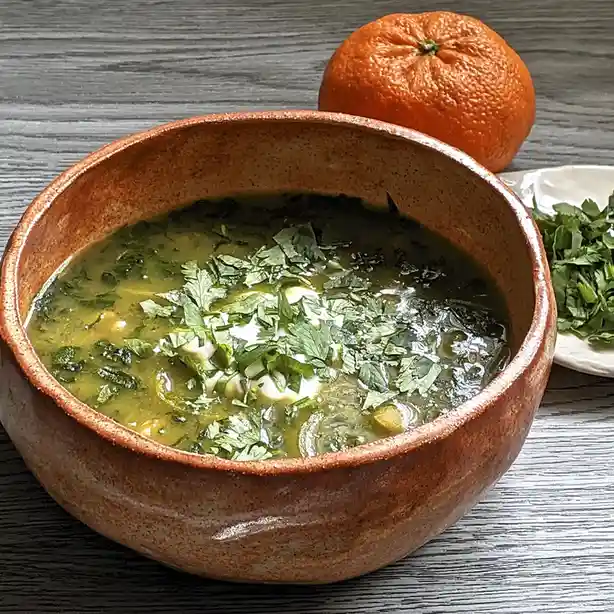 A rustic brown ceramic bowl filled with a green, herb-rich soup topped with dollops of white cream and chopped green herbs…
