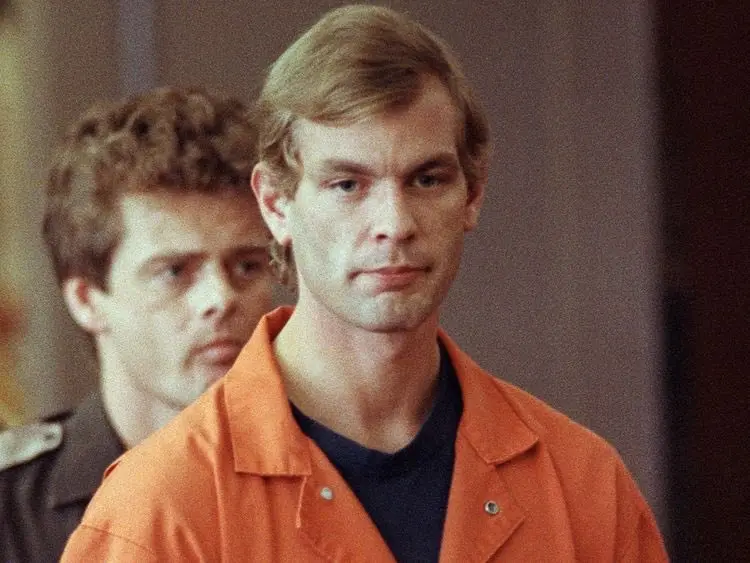 A man in an orange prison jumpsuit stares blankly ahead with a deadpan expression, while another man in a dark uniform…