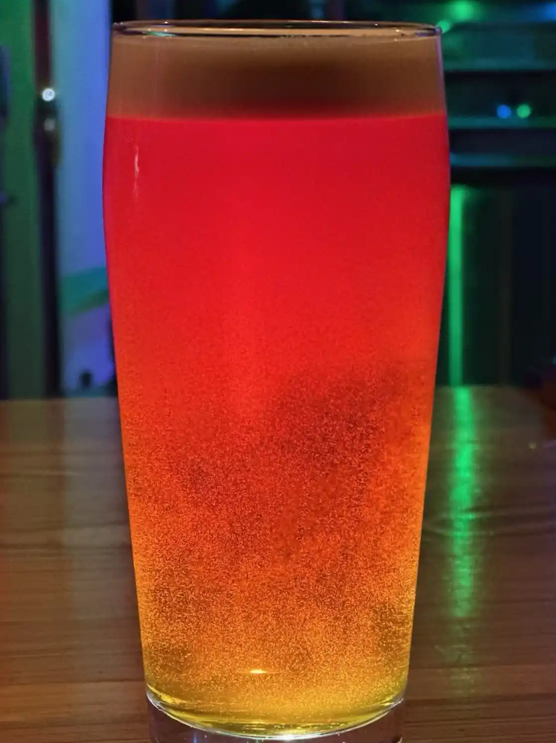 A tall glass of beer sits on a wooden table, its liquid displaying a striking vertical gradient from deep red at the top to…