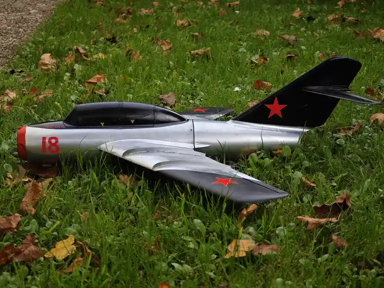 A silver model airplane with red stars and the number 18 on its fuselage lies on green grass scattered with brown leaves…