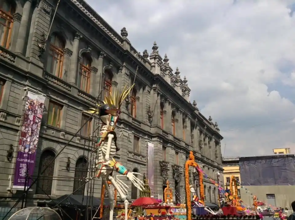 A large, ornate skeleton figure with a headdress and colorful decorations stands on a parade float in front of a grand, grey…