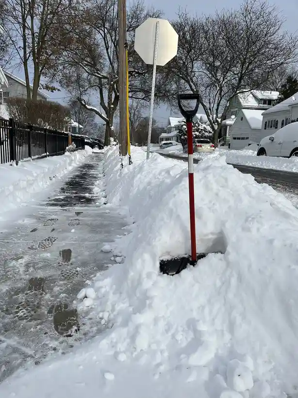 A red-handled snow shovel is stuck upright in a large pile of snow beside a cleared sidewalk, with a stop sign visible…