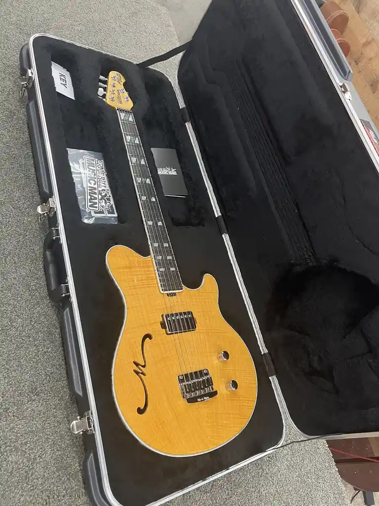 A light-wood, semi-hollow body electric guitar with a 'M' logo rests inside an open black hardshell case lined with black…