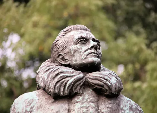 A close-up of a bronze statue of a man with a fur collar, looking upward and to the side. The statue has a weathered patina…