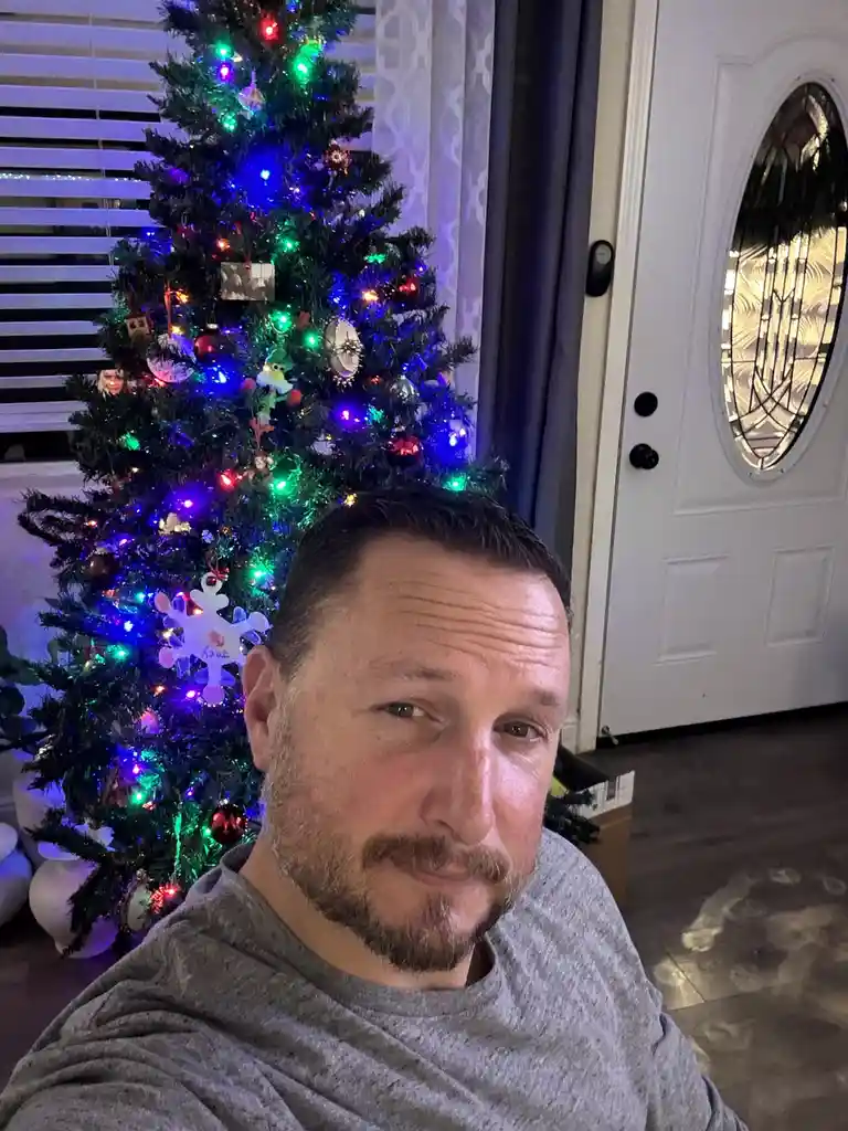A man with a beard and gray shirt takes a selfie in front of a brightly lit, decorated christmas tree with colorful lights…