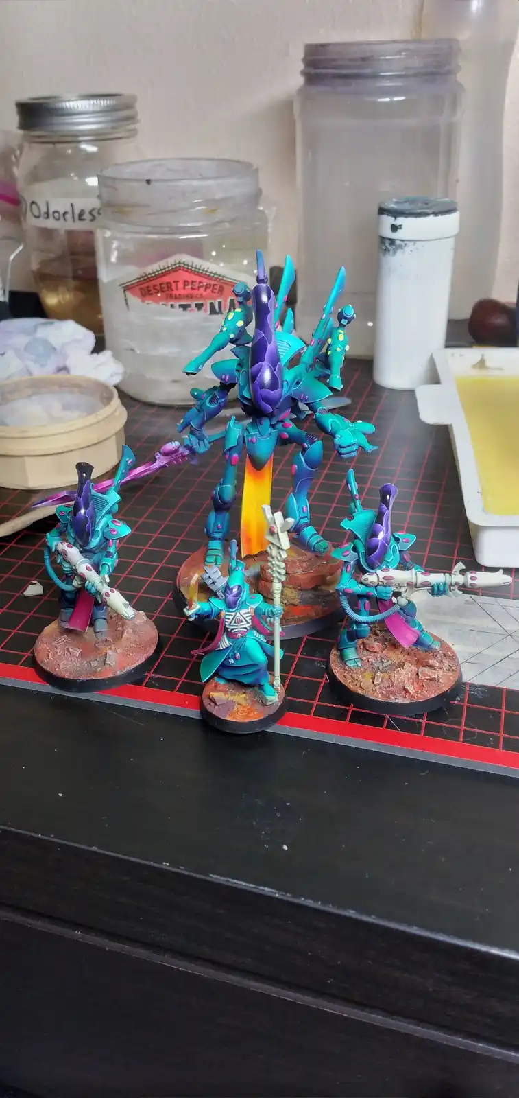 Four intricately painted fantasy miniatures, primarily in vibrant teal and purple with pink accents, stand on a red-grid…