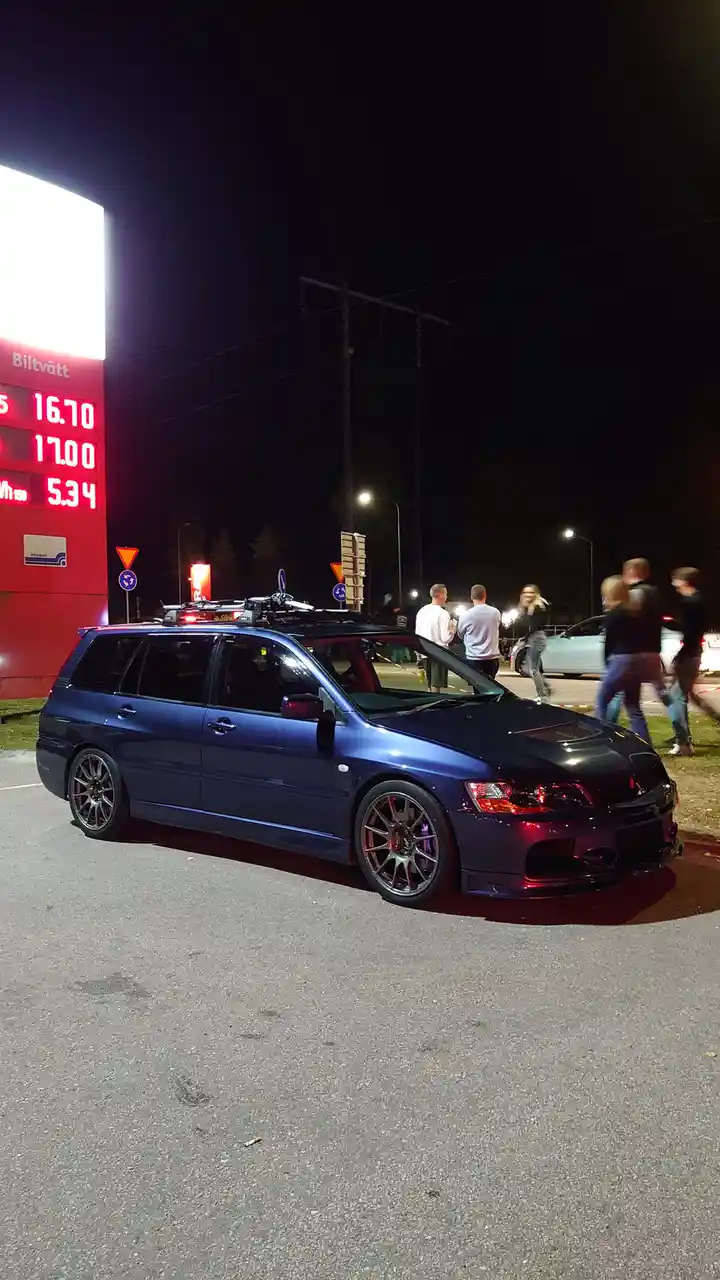 A dark blue Mitsubishi Lancer Evolution station wagon is parked at a Valvoline gas station at night, with a red digital sign…