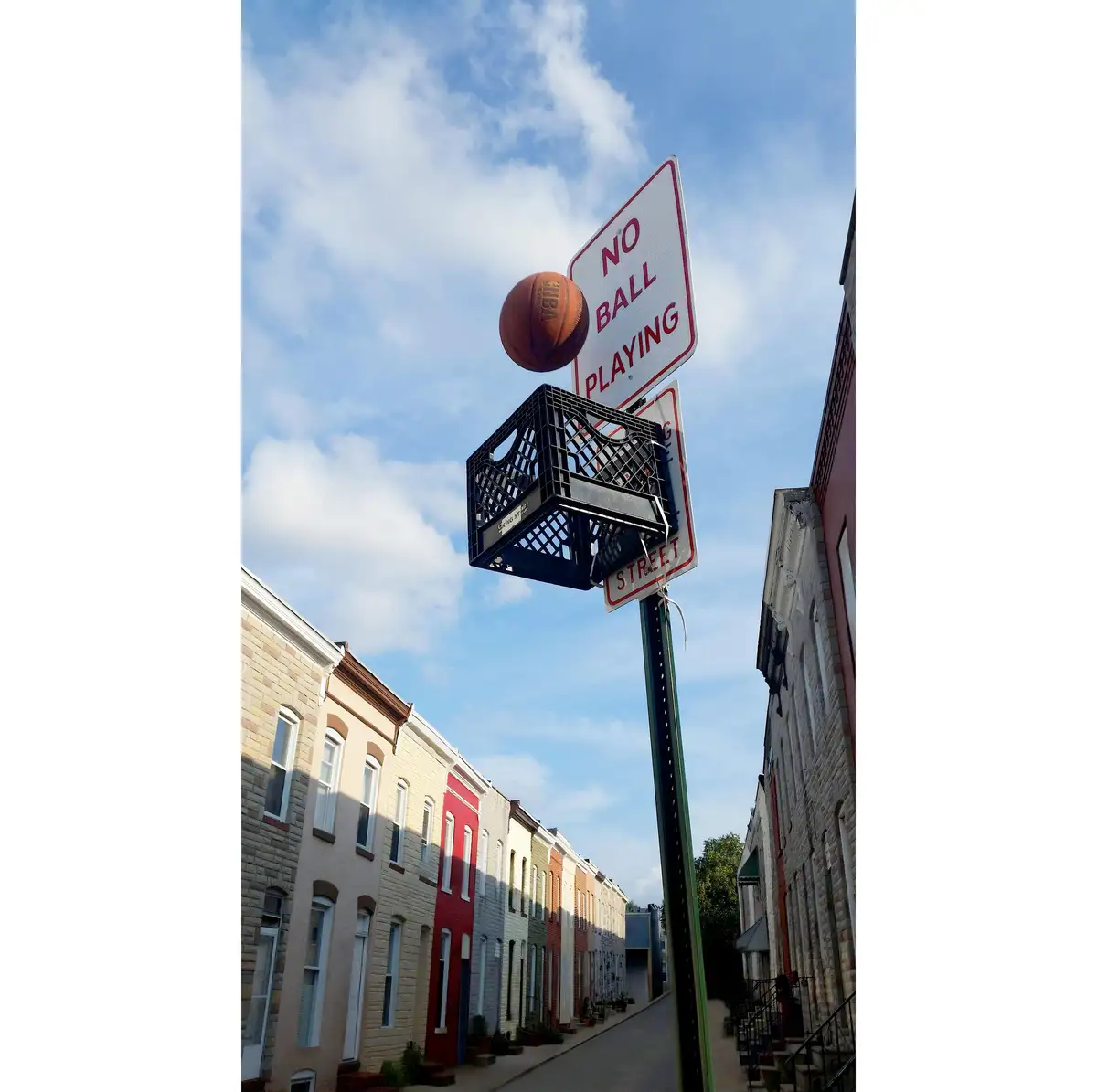 A basketball is suspended in mid-air just above a makeshift hoop made from a black plastic milk crate mounted on a pole with…