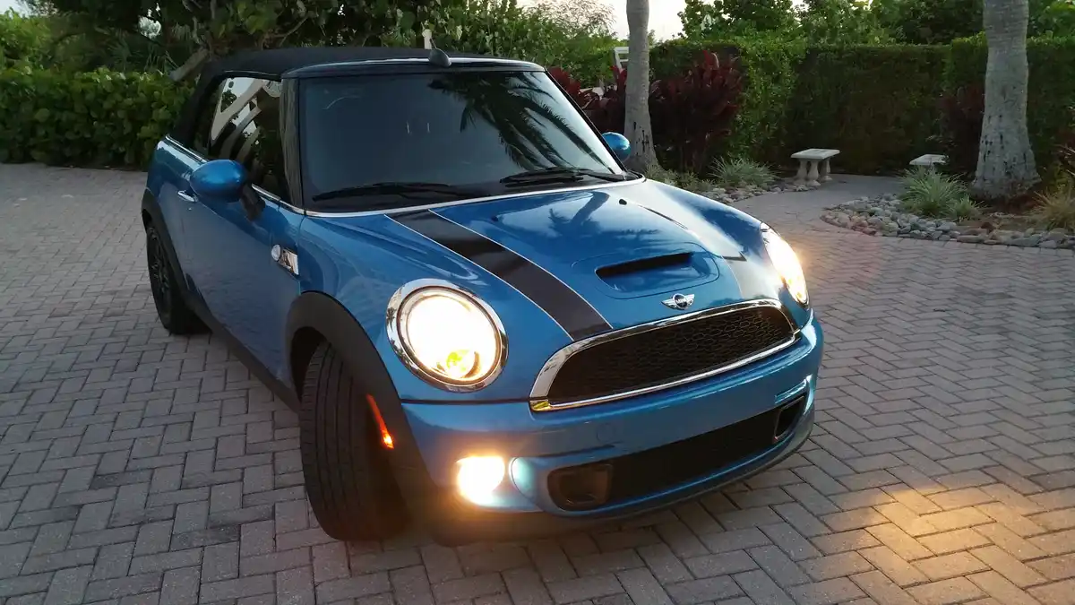 A blue Mini Cooper convertible with its headlights and fog lights on, parked on a brick driveway surrounded by lush greenery…
