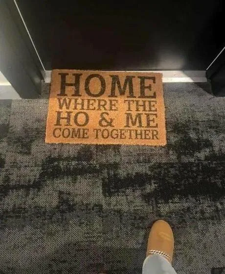 A close-up photo shows a person's foot in a tan slipper standing on a dark patterned carpet, facing a doormat that reads…