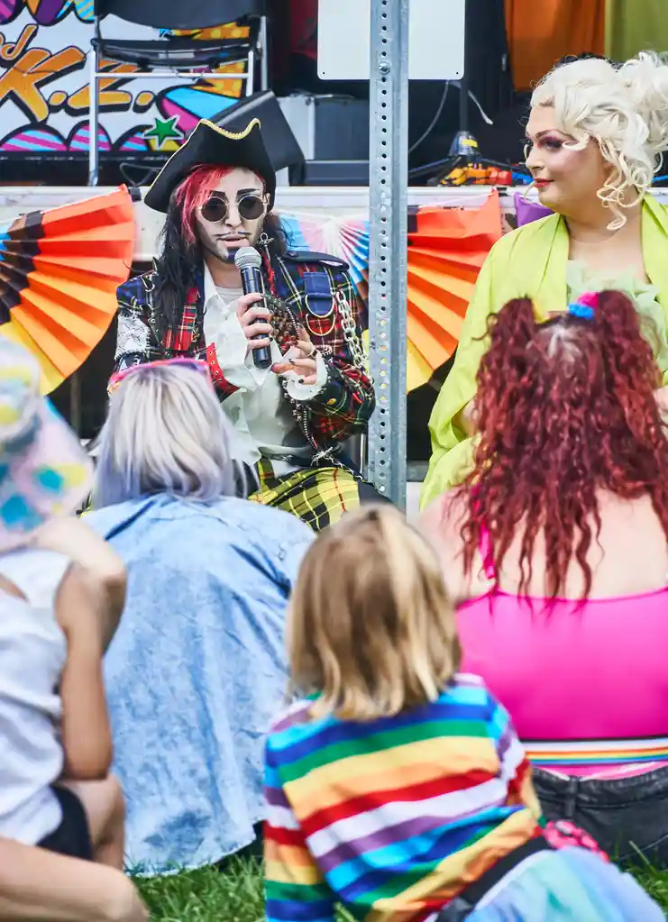 A person dressed as a pirate with a kilt, sunglasses, and a pirate hat holds a microphone while speaking to an audience at…