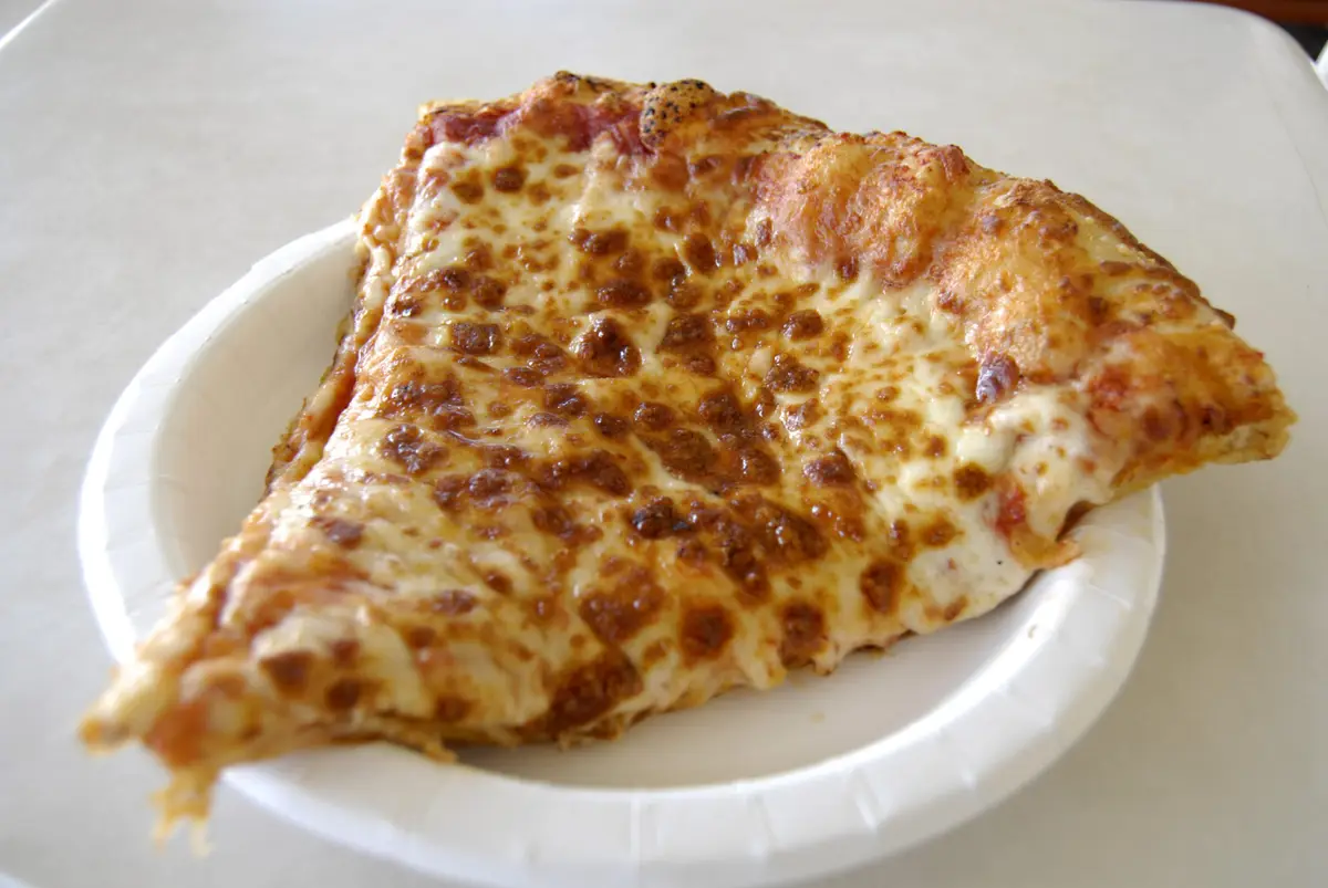 A single slice of cheese pizza with golden-brown, bubbly melted cheese sits on a white paper plate. The slice is thick and…