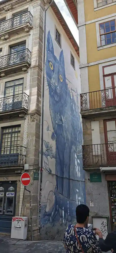 A large blue cat mural painted on the side of a building in a European city, with a person in the foreground looking at it…
