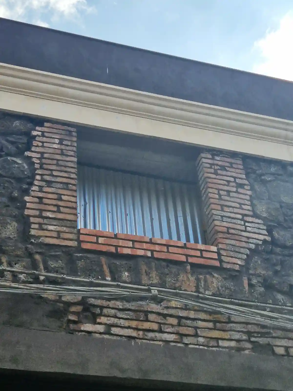 A close-up of a window on a weathered brick and stone building, framed by bricks and topped with a white molding, with…