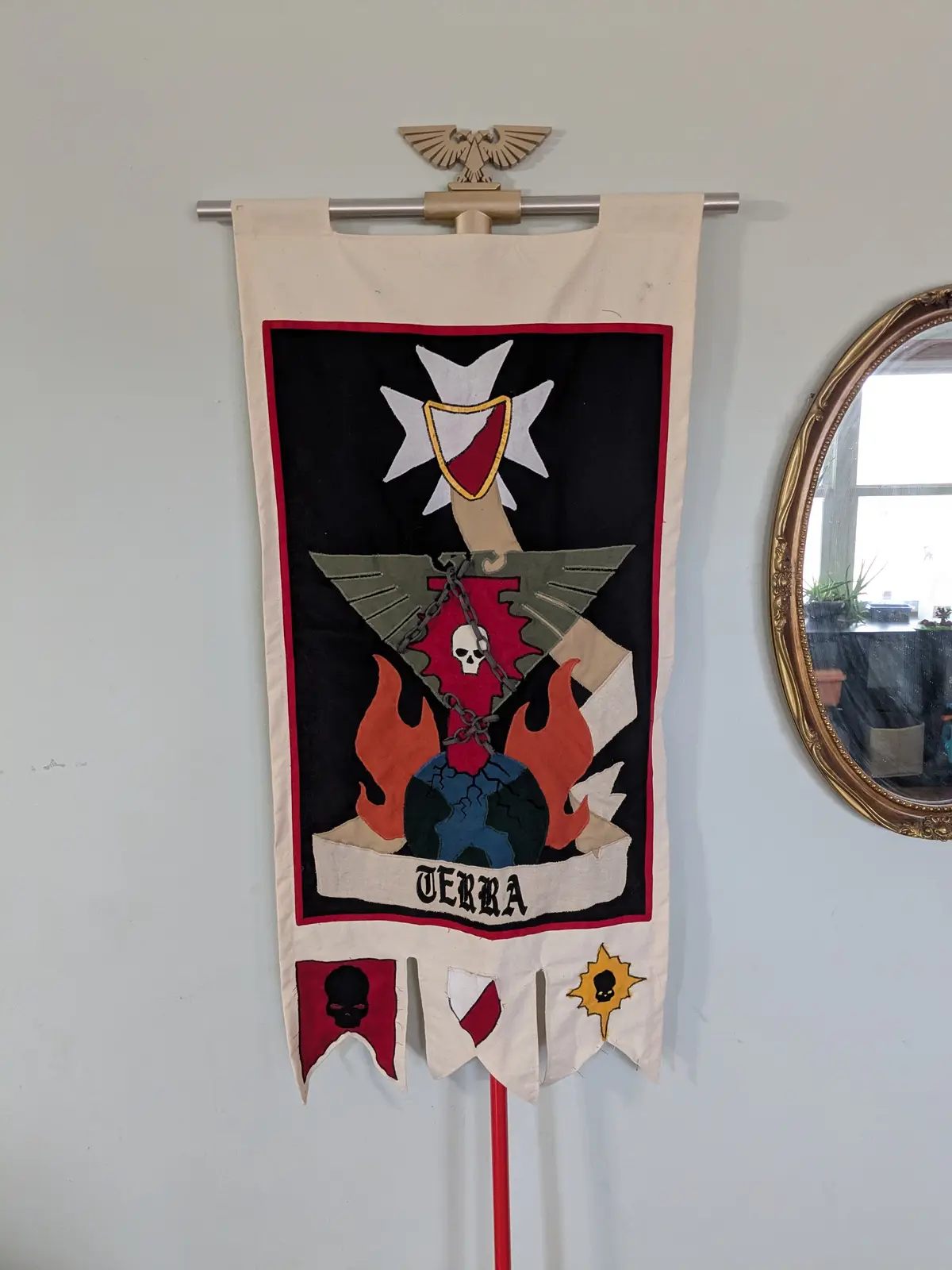 A handcrafted banner hangs on a wall, featuring a black field with a white eight-pointed star, a red shield, a skull on a…