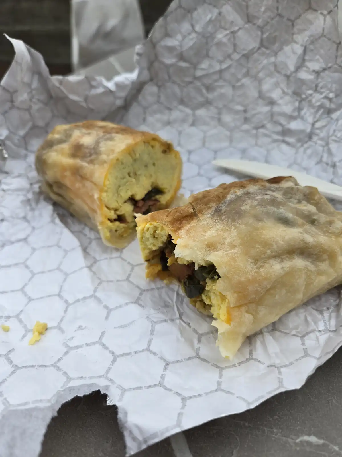 A close-up photo shows a breakfast burrito, cut in half, resting on hexagonal-patterned paper. The burrito has a…