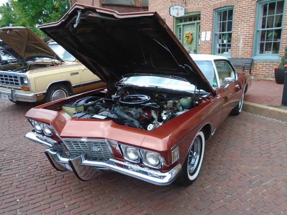 A shiny, rust-colored classic American car with its hood open, revealing a detailed engine, parked on a brick street in…