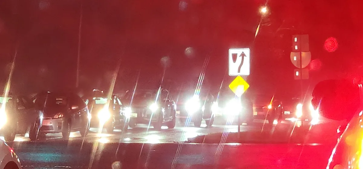 A nighttime traffic scene viewed from inside a car, showing multiple vehicles stopped with headlights on, a road sign…