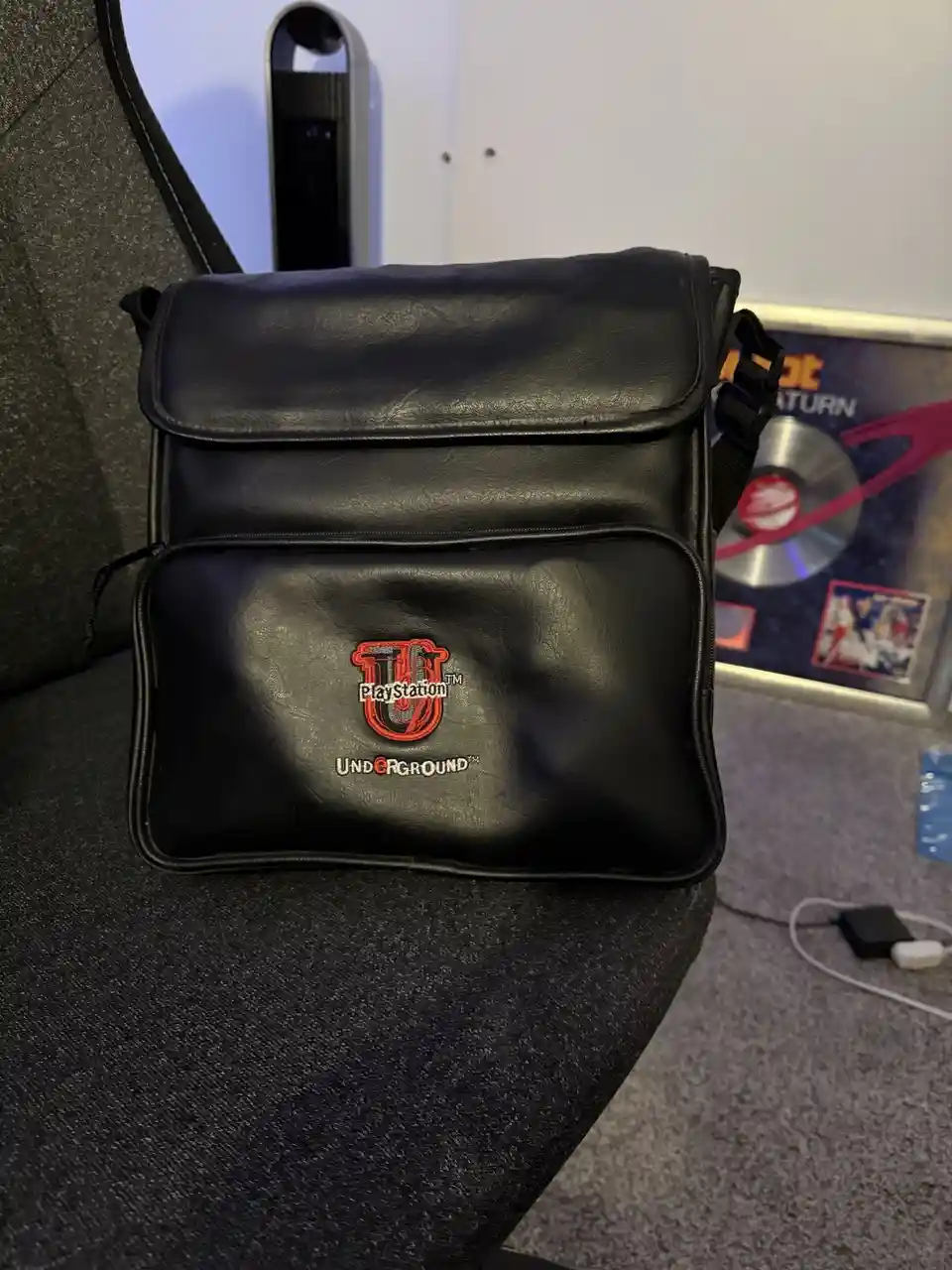 A black leather messenger bag with a red and white embroidered 'PlayStation Underground' logo sits on a dark gray chair. In…