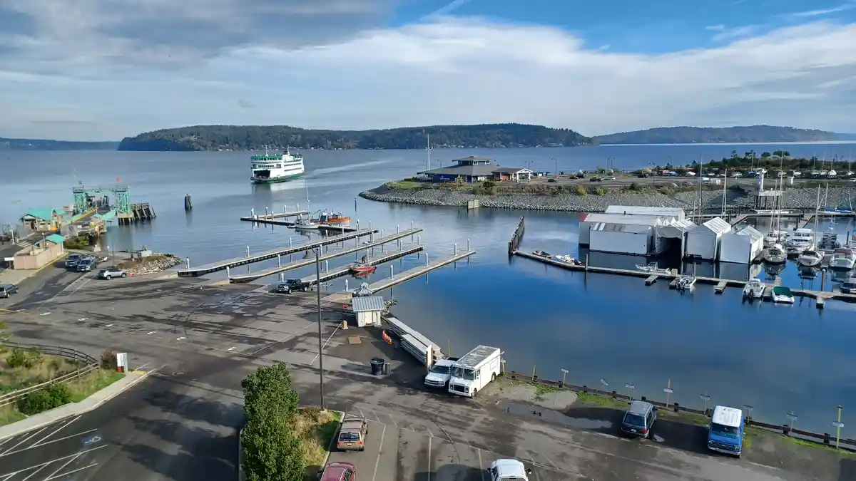 An aerial view of a harbor with docks, boats, a ferry, and parked vehicles under a partly cloudy sky. Overlaid text reads…