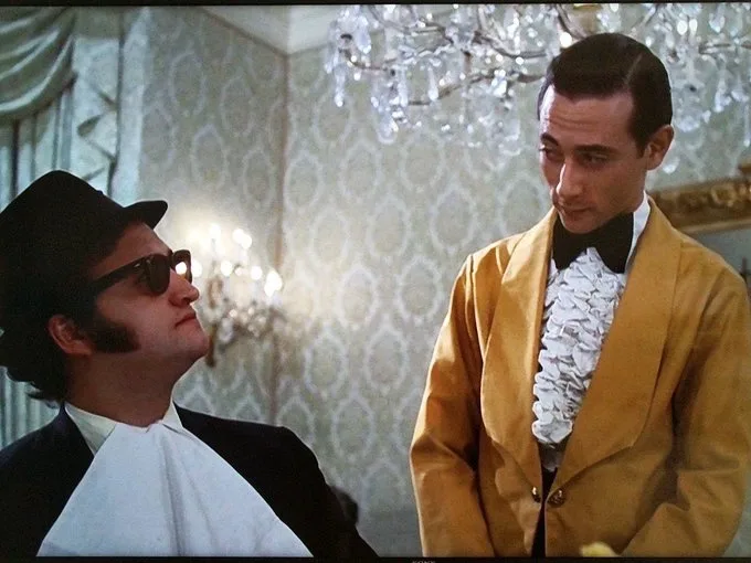 A man in a black suit, white collar, and sunglasses looks up confidently at another man in a yellow tuxedo with a white…